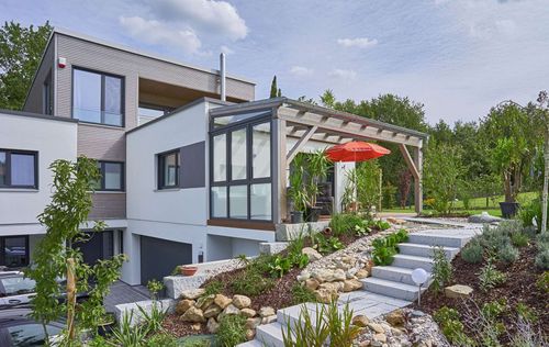 Die obere Terrasse des modernen Holzhauses im Bauhausstil mit drei Gebäudetrakten, zwei davon mit mineralischem Außenputz und Flachdach, einer mit Holzfassade aus Fichtenholzbohlen und Pultdach