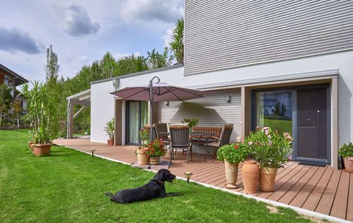 Die untere Terrasse im Garten des modernen Holzhauses im Bauhausstil mit drei Gebäudetrakten, zwei davon mit mineralischem Außenputz und Flachdach, einer mit Holzfassade aus Fichtenholzbohlen und Pultdach